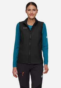 Eine Frau trägt eine schwarze Mammut-Weste über einem türkisfarbenen Langarmshirt und schwarzen Hosen mit orangefarbener Zipperlasche und steht vor einem schlichten Hintergrund.