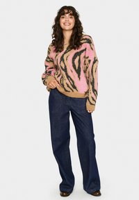 Maglione a maglia beige con motivo astratto rosa e nero; abbinato a pantaloni a gamba larga in denim scuro; la modella indossa scarpe marroni.