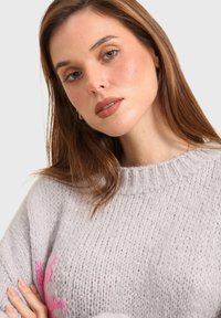 Pull en tricot gris clair avec un col côtelé et un motif floral rose. La texture semble douce et épaisse, mettant en avant des matériaux confortables.
