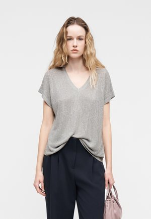 Hullámos szőke hajú nő ezüstszínű, rövid ujjú, V-nyakú kötött felsőt, fekete, magas derekú nadrágot visel, és egy kis mályva színű kézitáskát tart a kezében.