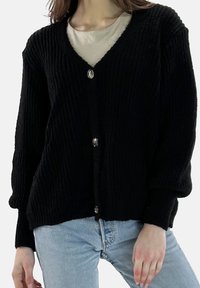 Zwarte gebreide cardigan met V-hals, twee zilveren knopen en een geribbelde structuur. Lange mouwen en een ontspannen pasvorm, gecombineerd met lichtblauwe spijkerbroek.