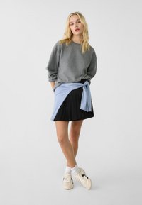Grau Sweatshirt, hellblauer Wickelrock zu einem schwarzen Faltenrock, kombiniert mit weißen Socken und cremefarbenen Sneakers mit schwarzen Akzenten.