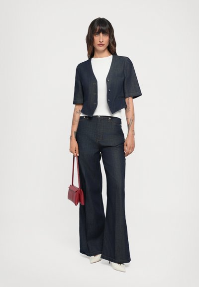Giacca cropped in denim scuro con maniche corte, abbinata a una t-shirt bianca e pantaloni flared in denim scuro. Accessoriata con una borsa rossa.