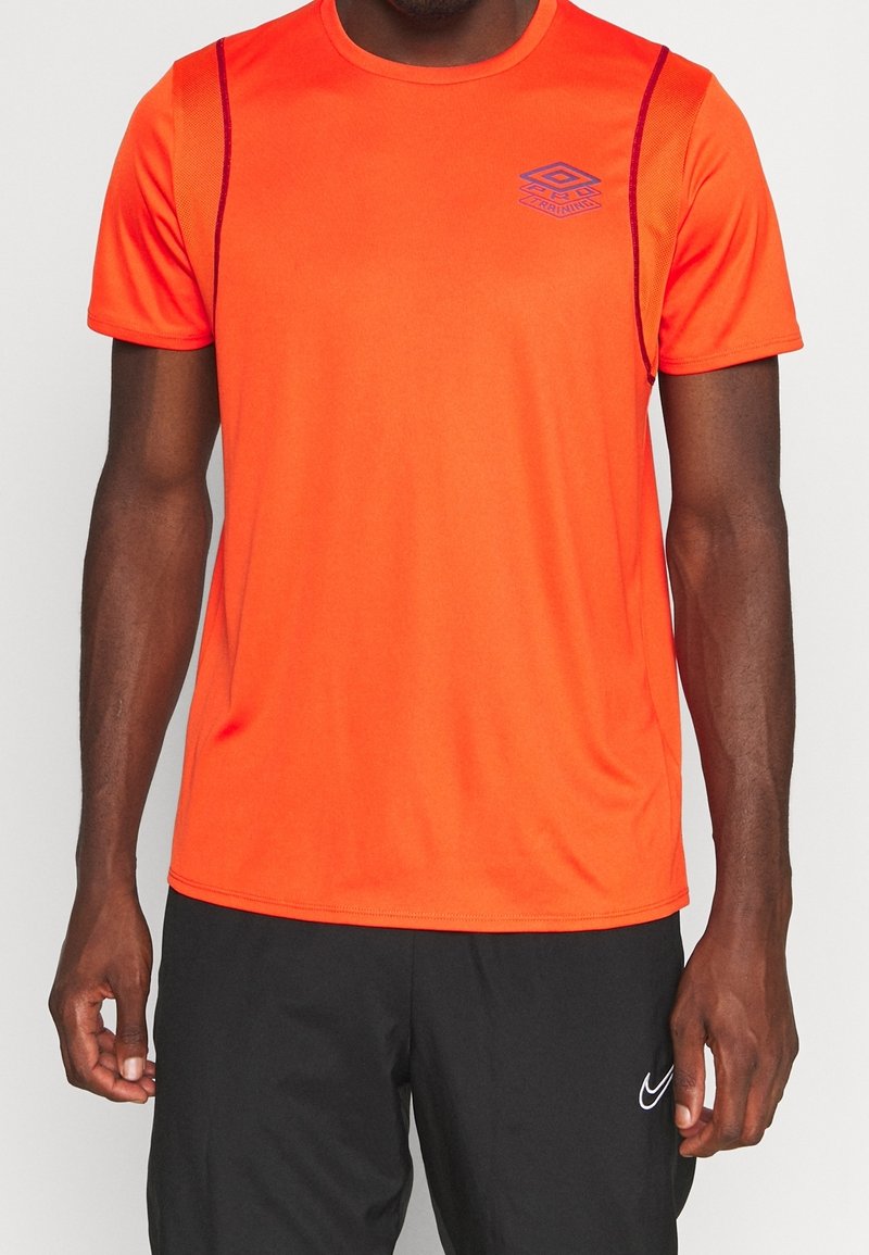 Homme portant un t-shirt de sport orange vif à manches courtes avec un petit logo et un short de sport noir avec un logo swoosh blanc sur la cuisse gauche.