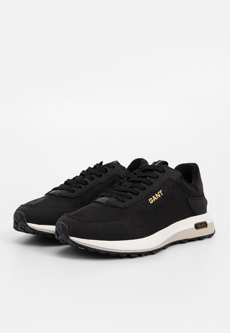 Zapatillas deportivas negras hechas de tela con una superficie texturada. Cuentan con cordones, un logo dorado y una suela en blanco y negro con un patrón rugoso.