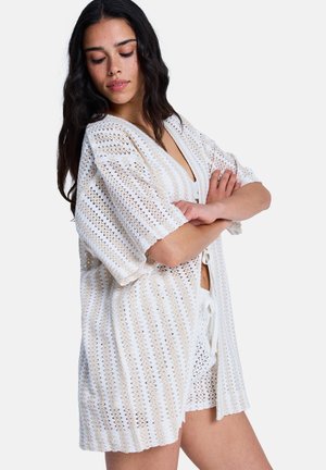 Cardigan en maille ouverte blanc avec une coupe ample, manches courtes, col en V profond et motif texturé. Porté avec un short assorti.