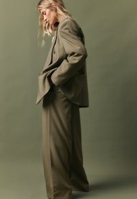 Pārmērīga olīvzaļā žakete ar vienu pogu, kombinēta ar platām biksēm. Audumam ir gluda tekstūra. Vienkāršs dizains.