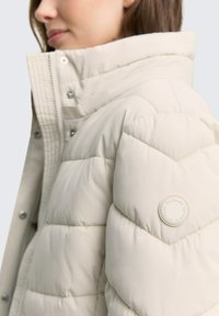 Chaqueta acolchada beige claro con acolchado en forma de chevrón, cuello alto y cierres a presión. Presenta un parche de logo redondo en la manga. Textura suave.