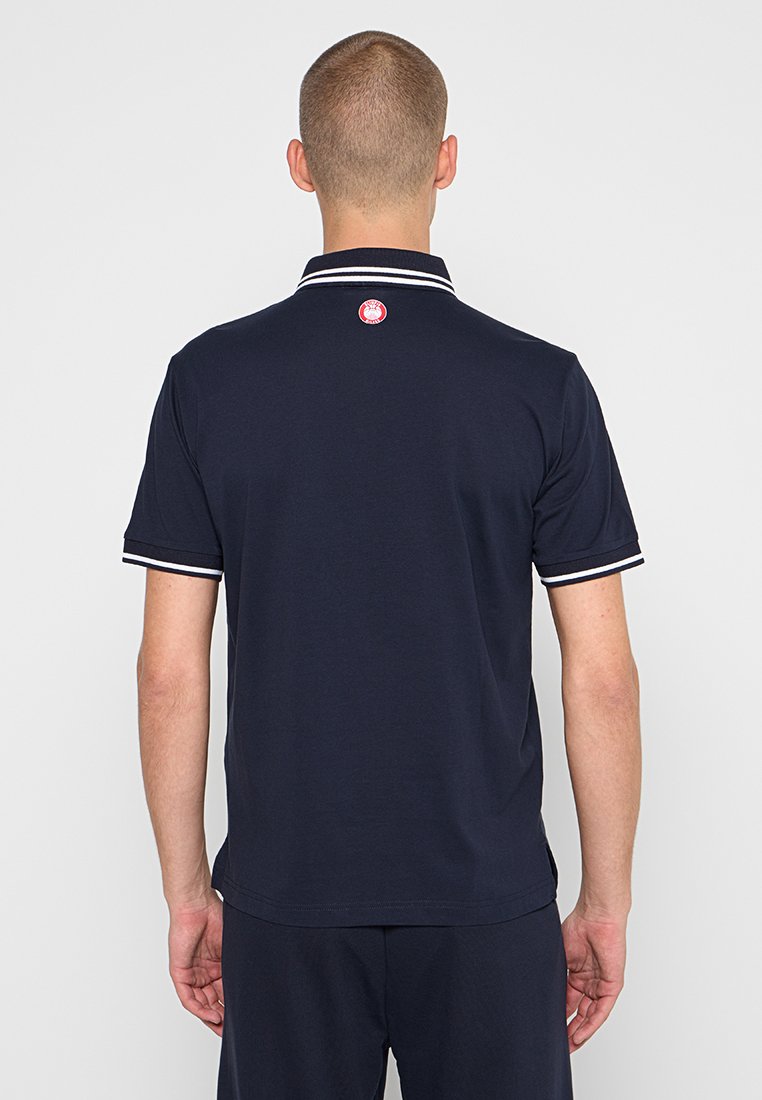 Granatowa koszulka polo z kołnierzykiem. Białe akcenty na kołnierzyku i rękawach. Posiada okrągłe logo na górnej części pleców. Wykonana z bawełnianego materiału.