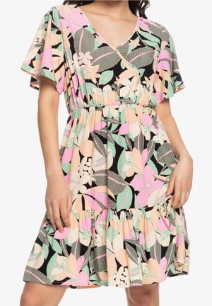Femme portant une robe portefeuille fleurie avec des feuilles et des fleurs roses, vertes et beiges, avec des manches courtes volantées et un ourlet à volants.