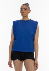 Blå ærmeløs top med rund hals og bred skulderdesign, lavet af et glat stof. Kombineret med sorte slidte shorts.