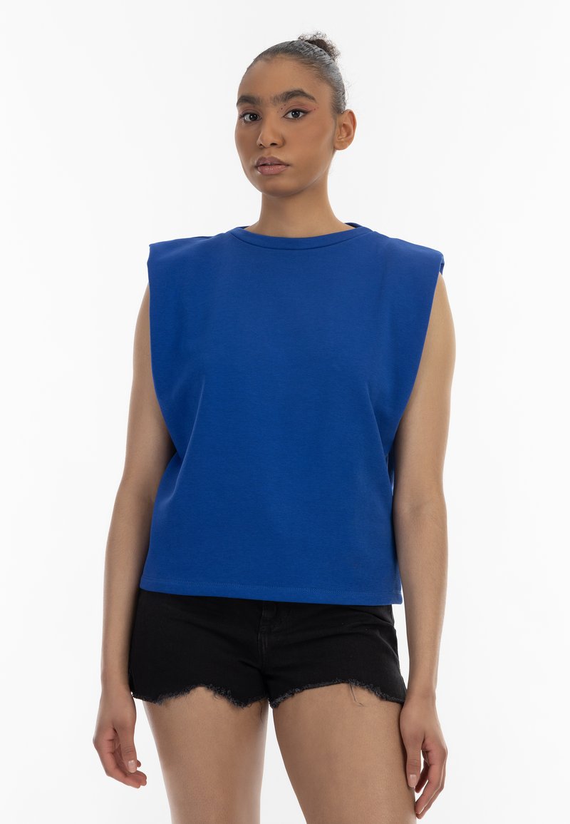 Blauwe mouwloze top met een ronde hals en een breed schouderontwerp, gemaakt van een gladde stof. Gecombineerd met zwarte uitgerafelde shorts.