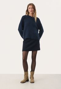 Pull en mailles bleu marine avec un motif en diamant texturé, associé à une mini jupe bleu marine et des bottines en suède beige à talon avec boucle sur le côté.
