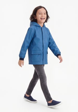 Barn går og smiler, iført en blå hættetrøje, grå jeans og navy slip-on sko mod en hvid baggrund.