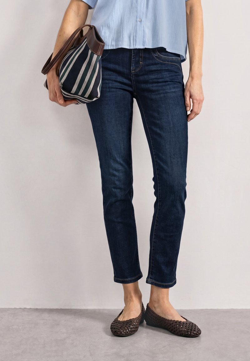 Femme portant un jean slim bleu foncé, des ballerines tressées marron, une chemise bleu clair, tenant un sac rayé avec des poignées en cuir dans la main gauche.