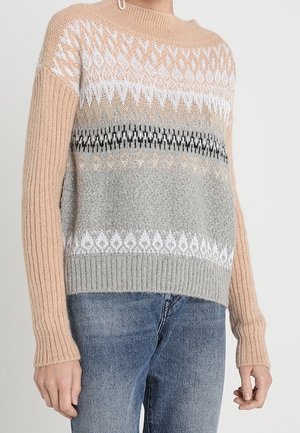 Pull en tricot avec manches beiges et corps à motifs en beige, blanc, gris et noir, associé à un jean bleu sur une personne debout.