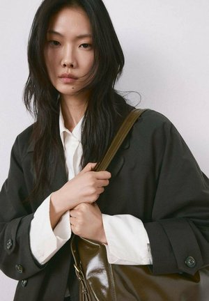 Joven con cabello largo y oscuro, vestida con un abrigo negro y una camisa blanca, sosteniendo un bolso grande de cuero color verde oliva.