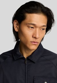 Giacca a zip blu navy con colletto, dotata di tasca sul petto e tessuto liscio e testurizzato. Design aderente con accenti sottili.