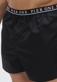 Shorts boxer noirs en tissu lisse, avec une ceinture noire portant le texte blanc "PIER ONE" et deux boutons à l'avant.