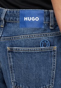Zilas džinsa bikses ar taisnstūrveida zilu ādas plāksteri ar uzrakstu "HUGO", redzamām šuvēm un mazu apaļu logotipu pie aizmugurējā kabatas.
