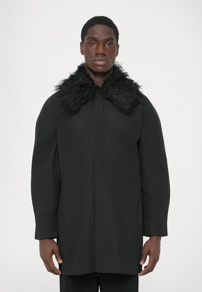 Hombre vestido con un largo abrigo negro con un gran y esponjoso cuello de piel negra, de pie contra un fondo blanco liso.