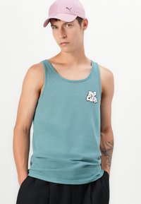 Mladic koji nosi tirkiznu majicu bez rukava s logom "Rip Curl", ružičastu kapu i crne hlače, stoji s rukama u džepovima.
