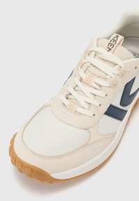 Baskets de sport beige et marine avec une tige synthétique lisse, un système de laçage, un col rembourré et une semelle en caoutchouc texturé.