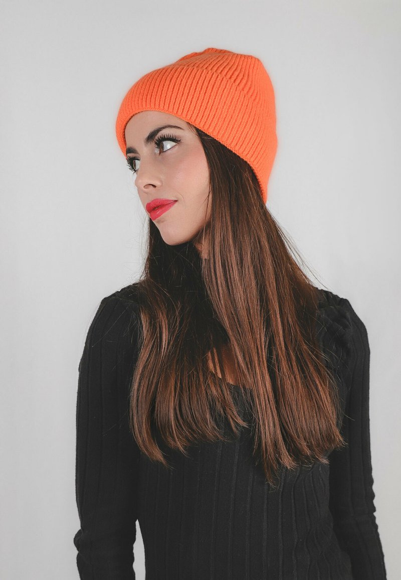 Beanie arancione a righe indossato da un modello con lunghi capelli castani, abbinato a un top nero a righe, su uno sfondo bianco semplice.