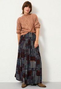 Ein Kabelstrickpullover in hellrosa, kombiniert mit einem bodenlangen gemusterten Rock in Dunkelrot, Marineblau und Creme. Braune Stiefeletten runden den Look ab.