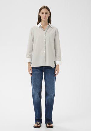 Woman with straight brown hair wearing a beige button-up shirt, blue jeans, and black sandals standing against a plain white background.