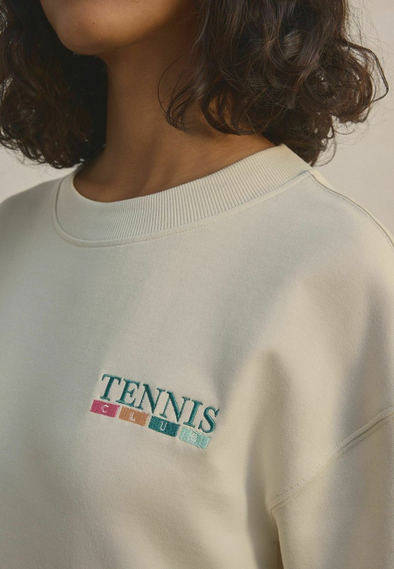 Kremowy sweatshirt z prążkowanym dekoltem, ozdobiony haftowanym napisem "TENNIS CLUB" w akcentach w kolorach zielonym, różowym i niebieskim.