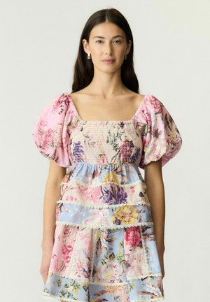 Kvinde med langt mørkt hår iført en blomsterprintet, lagdelt kjole med puffede korte ærmer og firkantet udskæring mod en ensfarvet lys baggrund.