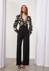 Pantalones de pierna ancha negros combinados con una blusa floral negra y dorada con mangas largas y un escote en V profundo. Texturas suaves, diseño elegante.