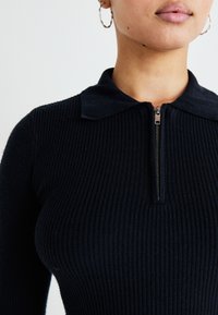 Black ribbed top with a zippered collar. Features textured fabric and a fitted silhouette, showcasing a close-up view of the neckline.