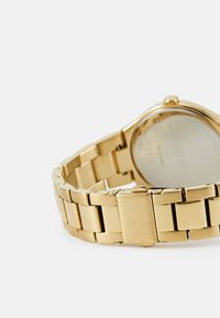 Reloj de metal dorado con una pulsera de eslabones, caja redonda y un acabado pulido. Parte trasera marcada con "Calvin Klein" y detalles técnicos.