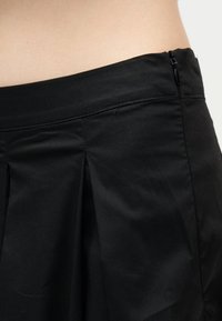 Μαύρη ψηλόμεση φούστα με πιέτες στο μπροστινό μέρος και πλευρικό φερμουάρ, που αποκαλύπτει μέρος της γυμνής μέσης πάνω σε απλό φόντο.