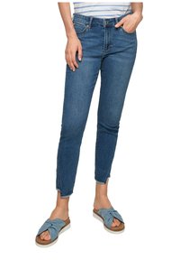 Blå denimjeans med slim fit, ankellängd och lätt slitning vid fållen, tillsammans med ljusblå knut-sandaler.