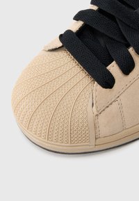 Hnedý semišový topánka s textúrovanou gumovou špičkou s geometrickým vzorom, čiernymi šnúrkami a hladkou čiernou vnútornou výstelkou.