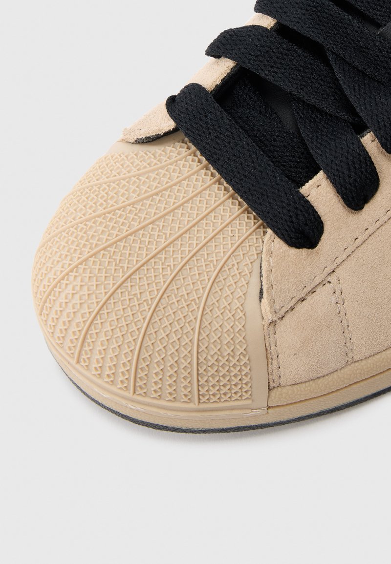 Hellbrauner Wildlederschuh mit einer strukturierten Gummikappe an der Spitze, die ein geometrisches Muster aufweist, schwarzen Schnürsenkeln und einem glatten, schwarzen Innenfutter.