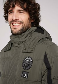 Chaqueta acolchada verde oliva con cuello alto, botones a presión y acentos negros. Presenta parches con logotipos de la marca en el pecho.