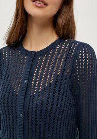 Mornarska pletena jopica z odprtim vzorcem, okroglo ovratnikom, zapiranjem na gumbe in teksturiranimi rebrastimi detajli. Vidni gumbi in šivani detajli.