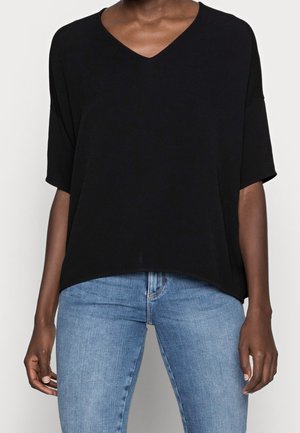 Personne vêtue d'un haut ample noir à col en V et manches mi-longues, associé à un jean bleu clair ajusté, se tenant devant un fond uni.