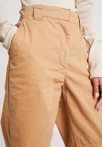 Pantalons larges beiges en coton, dotés d'une taille haute, de poches latérales et d'une chaîne en métal visible sur la boucle de ceinture.