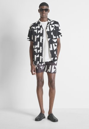 Hombre con camisa de manga corta con estampado geométrico en blanco y negro y pantalones cortos a juego, camiseta blanca, zapatos negros tipo slip-on y gafas de sol.