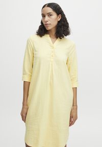 Robe jaune à boutons avec des manches trois-quarts, en tissu léger. Présente un décolleté en V et un design plissé pour une texture supplémentaire.