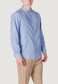 Camicia button-down azzurro chiaro a maniche lunghe con colletto a fascia. Presenta bottoni a contrasto e una leggera trama. Abbinata a pantaloni beige a righe.
