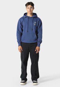 Felpa blu in cotone con tasca frontale, cappuccio regolabile con cordino e dettagli ricamati. Indossata con pantaloni neri e scarpe da ginnastica.