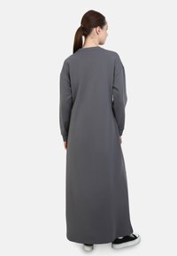 Langes graues Sweatshirtkleid aus weichem Stoff, mit Rundhalsausschnitt, langen Ärmeln und seitlichen Schlitzen, kombiniert mit schwarzen Sneakers.