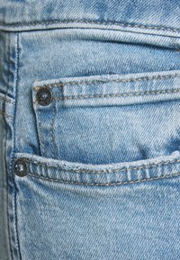 Jasnoniebieskie dżinsy z denimu z tylną kieszenią zapinaną na ciemny guzik. Tkanina ma lekko wyblakłą fakturę z widocznymi detalami przeszyć.