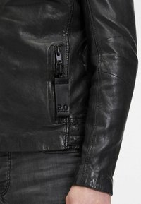 Giacca in pelle nera con tasca zip e etichetta logo embosser. Il materiale ha una consistenza liscia con dettagli di cucitura sottili.